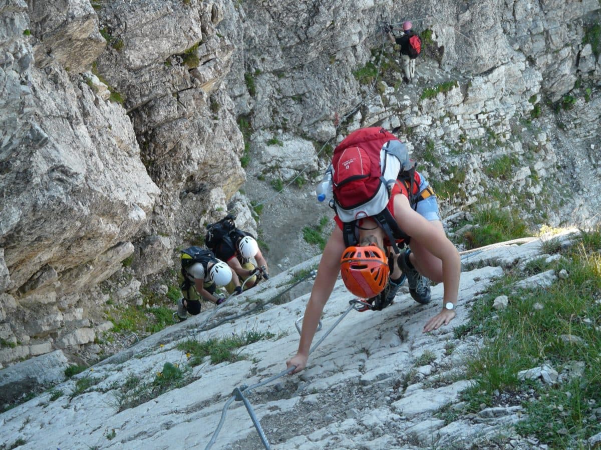 Nell’immagine una scena di alpinismo
foto a colori, parete in verticale e la visuale dall'alto,  in primo piano il primo alpinista, si vedono la testa protetta dall'elmetto rosso, le spalle con lo zainetto bianco e rosso, le scarpe da alta montagna e il moschettone collegato alla fune di acciaio di sicurezza ancorata a parete.  Dietro in secondo piano altri due compagni in cordata equipaggiati nello stesso modo. 
In lontananza un'altra persona – probabilmente un istruttore - che fissa o controlla la fune di sicurezza. 
Il messaggio sottinteso: La conoscenza dei pericoli e l’adozione delle cautele possibili sono necessari per minimizzare i rischi. 
 
