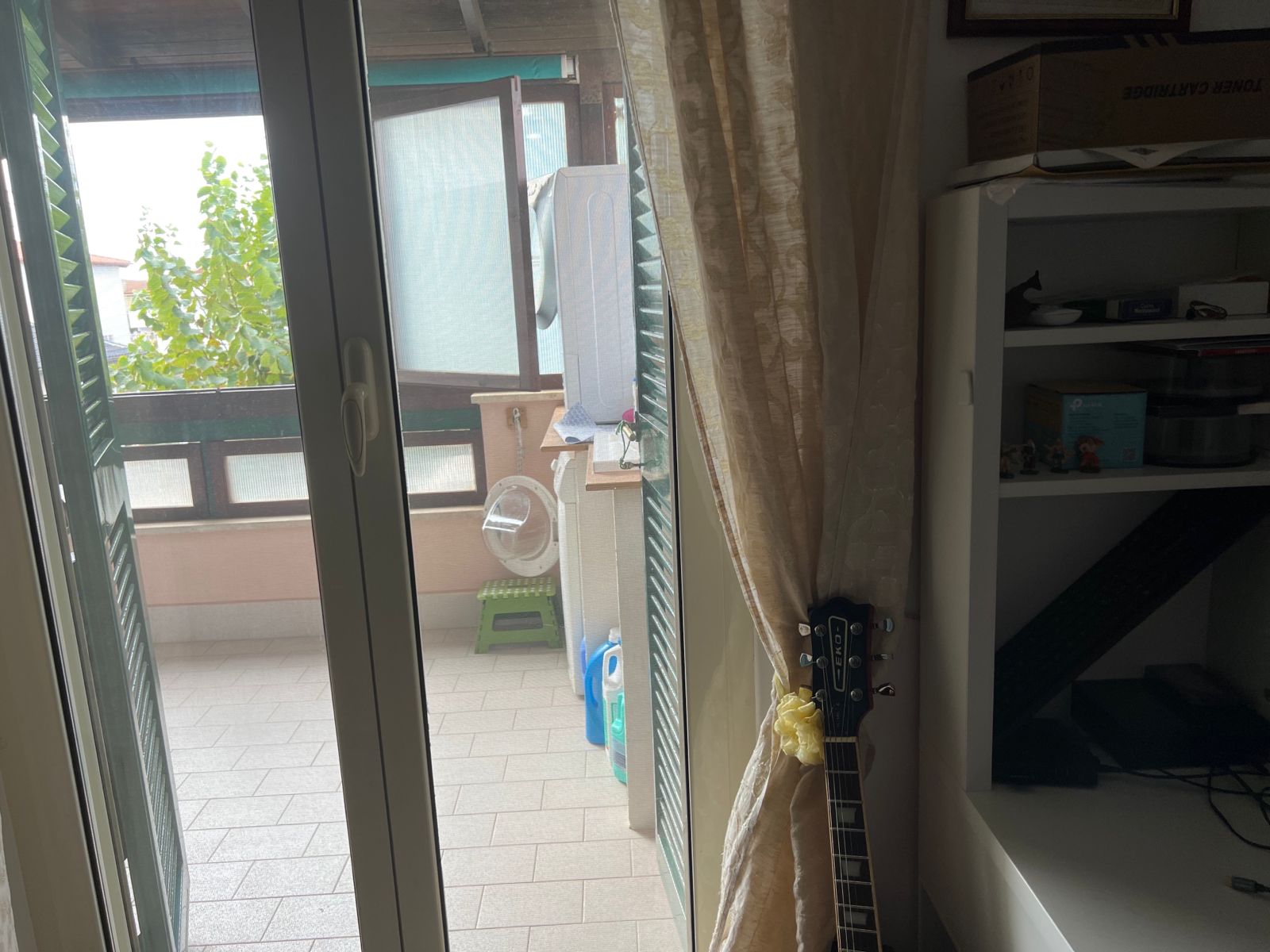 La foto mostra un angolo di una stanza con una porta-finestra aperta che conduce a un balcone. Sul balcone c'è un asse da stiro con una copertura bianca e un secchio verde. Ci sono anche alcune piante verdi visibili fuori. All'interno, accanto alla porta-finestra, c'è una tenda color crema. A destra della tenda, c'è una chitarra appoggiata al muro con un fiocco giallo legato vicino alla parte superiore del manico. Accanto alla chitarra c'è una libreria bianca con vari oggetti, tra cui scatole e un contenitore blu.